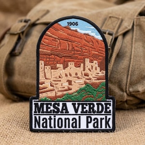 May include: An embroidered patch featuring a landscape of Mesa Verde National Park with cliff dwellings, red rock formations, and green vegetation. The top arch displays the year 1906, and the base features the text MESA VERDE National Park.