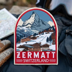 May include: An arched embroidered patch featuring a detailed landscape of the Matterhorn mountain in Zermatt, Switzerland. The design includes snow-covered peaks, pine trees, and wooden chalets, with the text ZERMATT SWITZERLAND in white on red.