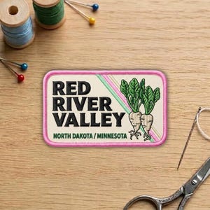 May include: Embroidered patch with the text "RED RIVER VALLEY" and "NORTH DAKOTA / MINNESOTA" in black. The patch features a pink border and a graphic of two beets with green leaves. Sewing supplies are in the background.