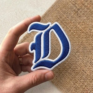 May include: Embroidered patch in the shape of the letter "D". The patch features a bold, blue gothic-style font with white accents and a white border. The patch is likely for clothing or accessories.