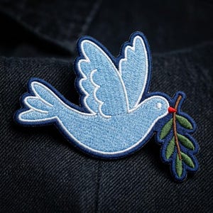May include: An embroidered patch featuring a light blue dove in flight with a dark blue border. The bird holds a green olive branch with a small red berry in its beak. The design is stitched with visible thread texture on a dark fabric surface.