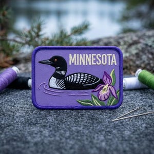May include: A rectangular embroidered patch with a purple background featuring a black and white common loon swimming on water, a purple lady slipper flower, and the word MINNESOTA in white capital letters with a dark purple stitched border.