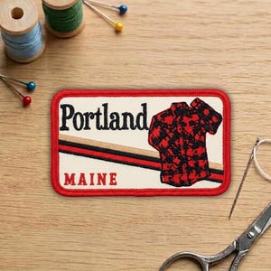 May include: A rectangular patch with a red border features the word "Portland" above "MAINE". A red and black plaid shirt graphic is on the right. The patch is on a wooden surface with sewing supplies.