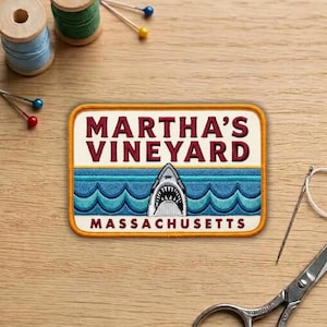 May include: Embroidered patch featuring the text "MARTHA'S VINEYARD MASSACHUSETTS" with a shark graphic. The patch has a rectangular shape with a cream-colored border, blue water, and a shark with an open mouth.