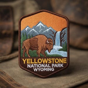 May include: A hexagonal embroidered patch featuring a brown bison standing in a green field. The background shows blue mountains, a waterfall, and an orange sky. The text reads YELLOWSTONE NATIONAL PARK WYOMING in yellow and white capital letters.