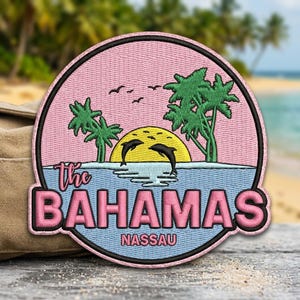 May include: A circular embroidered patch featuring a pink sky, a yellow sun, and two black dolphins leaping over blue water. Green palm trees frame the scene. The text reads "the BAHAMAS" in pink and "NASSAU" in smaller pink letters below.