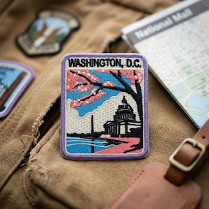 May include: A rectangular embroidered patch featuring the text WASHINGTON, D.C. at the top. The design depicts a black silhouette of the U.S. Capitol building and the Washington Monument against a blue sky, with pink cherry blossom branches.