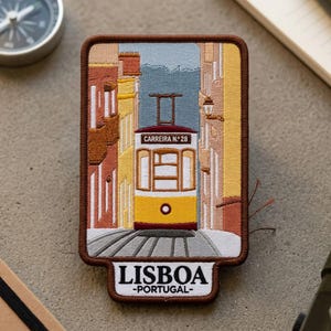 May include: A rectangular embroidered patch featuring a yellow Lisbon tram on tracks between buildings. The tram displays the text CARREIRA N. 28. Below the tram, the patch features the text LISBOA -PORTUGAL- in black letters on a white background.