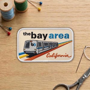 May include: Embroidered patch featuring a train with the text "the bay area" and "California". The patch has a cream background with a train design and colorful stripes. Additional sewing supplies are in the background.