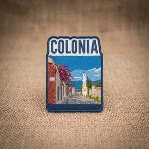 May include: A rectangular embroidered patch with a dark blue border featuring the word COLONIA in bold blue letters at the top. The design depicts a cobblestone street leading toward a white lighthouse, a brick building, and pink flowers.