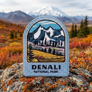 May include: An embroidered patch featuring a landscape of snow-capped mountains, evergreen trees, and a golden meadow. The patch has a light blue border and the text DENALI NATIONAL PARK in black capital letters at the bottom of the design.