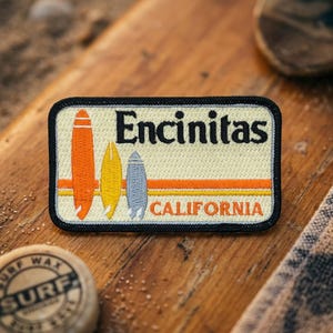 May include: Embroidered patch featuring three surfboards in orange, yellow, and blue. The patch has the word "Encinitas" above "California" in a retro design, with a black border.