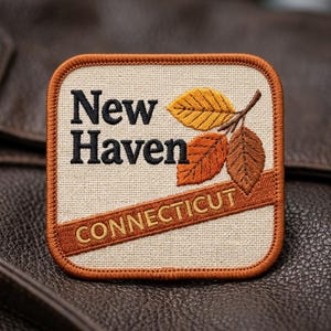 May include: A square embroidered patch with a cream-colored fabric background and a rust-orange border. The text New Haven is stitched in black, with Connecticut in gold on a diagonal stripe. Three autumn leaves in yellow, orange, and brown.