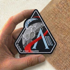 May include: Black, triangular patch featuring a detailed illustration of the moon, Earth, and a stylized letter "A". The patch includes the names "WISEMAN", "GLOVER", "KOCH", and "HANSEN". Red and blue accents enhance the design.