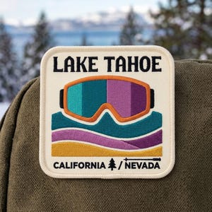May include: A square embroidered patch with a cream background featuring the text LAKE TAHOE at the top and CALIFORNIA / NEVADA at the bottom. The design includes a pair of orange-rimmed ski goggles above stylized teal, purple, and gold waves.