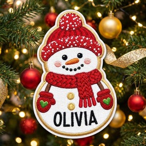 May include: A white snowman patch featuring a red knit hat with white dots, a red scarf, and red mittens with green hearts. The snowman has a carrot nose, black eyes, and a black mouth. The name OLIVIA is embroidered in black at the bottom.