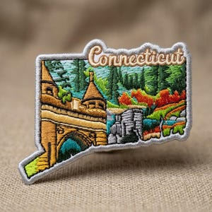 May include: An embroidered patch shaped like the state of Connecticut with a silver border. The design features a tan stone bridge, a dark blue archway, green pine trees, and autumn-colored foliage. The word Connecticut is stitched in tan.