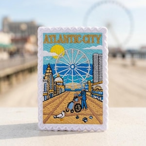 May include: A rectangular embroidered patch with a scalloped white border featuring the text ATLANTIC CITY in gold. The design depicts a boardwalk scene with a large Ferris wheel, a person in a wheelchair, a seagull, and a bright yellow sun.