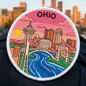May include: A circular embroidered patch featuring a skyline illustration with the text OHIO at the top. The design includes a tall tower, city buildings, a bridge, and a winding blue river against a pink and orange sunset sky with a yellow sun.