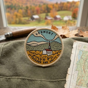 May include: A circular embroidered patch featuring a landscape with a white church, yellow trees, and blue mountains. The text VERMONT is arched at the top, and FREEDOM AND UNITY is arched at the bottom. The patch has a cream-colored border.