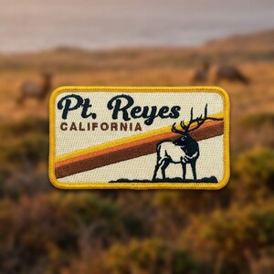 May include: A rectangular embroidered patch with a cream background and a yellow border. It features the black text Pt. Reyes CALIFORNIA, a diagonal orange and brown stripe, and a black silhouette of an elk standing on dark ground.