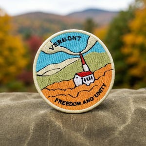 May include: A circular embroidered patch featuring a landscape with a white building with a red roof, green hills, and a blue sky. The text VERMONT is arched at the top, and the text FREEDOM AND UNITY is arched at the bottom in black thread.