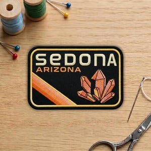May include: A black patch with the text "SEDONA ARIZONA" in cream-colored letters. The patch features an orange and yellow crystal design and a diagonal orange stripe. The patch is rectangular with rounded corners and a yellow border.