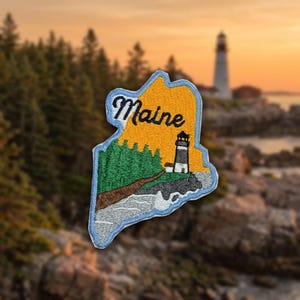 May include: An embroidered patch shaped like the state of Maine, featuring a yellow sky, green pine trees, a brown path, and a black and white lighthouse on a rocky gray coastline. The word Maine is stitched in black cursive script at the top.