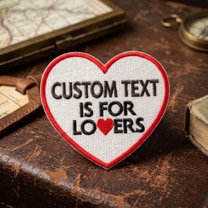 May include: A heart-shaped embroidered patch with a white background and a red border. The patch features the black embroidered text CUSTOM TEXT IS FOR LOVERS, with a small red heart replacing the letter V in the word LOVERS.