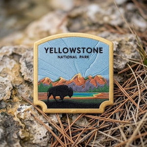 May include: A rectangular embroidered patch with a gold border featuring the text YELLOWSTONE NATIONAL PARK in black. The design depicts a black bison in the foreground, a turquoise pool, green pine trees, and orange mountains under a light blue sky.