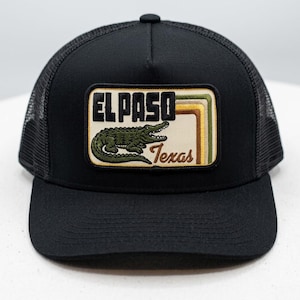 May include: Black trucker hat with a mesh back and a curved brim. The front features a rectangular patch with the text "EL PASO Texas" and an illustration of an alligator in green and brown.