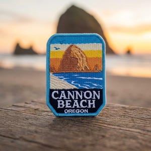 May include: A rectangular embroidered patch with rounded corners featuring a landscape of Cannon Beach, Oregon. The design shows a large brown rock formation, a sandy beach, blue ocean waves, and a sunset sky. The text reads CANNON BEACH OREGON.