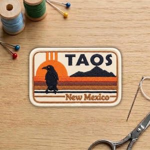 May include: Embroidered patch featuring a black raven in front of a sunset, with the text "TAOS New Mexico." The patch has a rectangular shape with a cream-colored border and is set against a wooden surface.