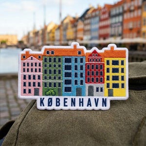 May include: An embroidered patch featuring a row of colorful buildings in pink, green, blue, red, and yellow. A small green bicycle is depicted on the green building. The text KØBENHAVN is embroidered in dark blue capital letters at the bottom.