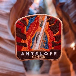 May include: A rectangular embroidered patch with rounded corners featuring a colorful depiction of Antelope Canyon. The design showcases red, orange, and blue rock formations with a central light beam. The text ANTELOPE CANYON is in white.