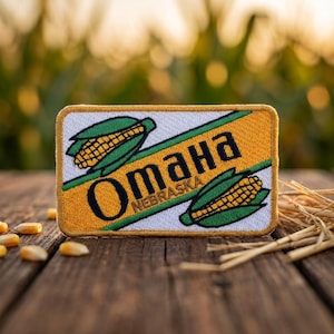 May include: A rectangular embroidered patch featuring a yellow and white background with two illustrations of corn cobs with green husks. The text OMAHA NEBRASKA is stitched in black across the center, framed by a gold-colored stitched border.