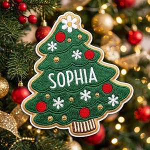 May include: A green embroidered Christmas tree patch featuring the name SOPHIA in white letters. The tree is decorated with red circular ornaments, small gold dots, white snowflake patterns, and a white flower at the top peak.