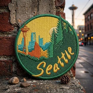 May include: A circular embroidered patch featuring a yellow sky, a stylized Seattle skyline with the Space Needle in orange and blue, and green pine trees. The word Seattle is stitched in yellow cursive script across the bottom of the patch.