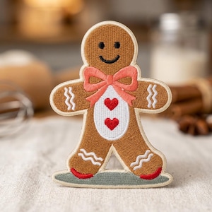 May include: A brown gingerbread person embroidered patch featuring a smiling face, a coral-colored bow at the neck, and a white belly panel with two red hearts. The arms and legs have white wavy line details, and the feet are accented with red.