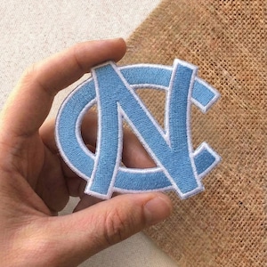May include: A light blue embroidered patch featuring the letters "NC" inside a light blue circle. The patch has a white border and is held against a tan fabric background.