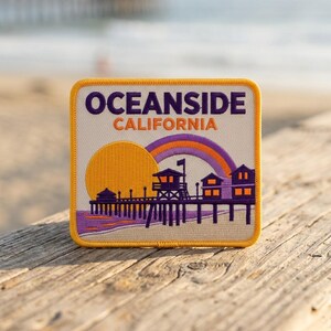 May include: A square embroidered patch featuring the text OCEANSIDE CALIFORNIA in dark purple and orange. The design depicts a yellow sun, a purple rainbow, and a dark purple pier with buildings over a sandy beach background with light colors.