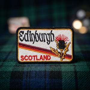 May include: A rectangular embroidered patch featuring the word Edinburgh in black script, the word SCOTLAND in red capital letters, and a stylized thistle flower in shades of red, black, and gold, all set against a white background with a black border.