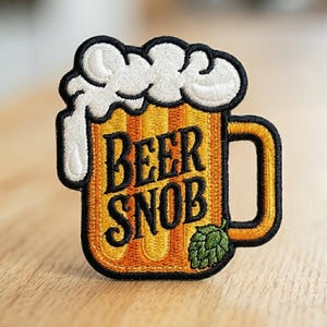 May include: An embroidered patch shaped like a beer mug with a yellow body, a white foamy top, and a black handle. The text BEER SNOB is stitched in black letters across the center, with a small green hop illustration at the bottom right corner.