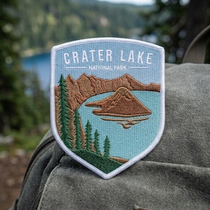 May include: A shield-shaped embroidered patch featuring a light blue lake, brown mountains, and green pine trees. The text CRATER LAKE NATIONAL PARK is stitched in white at the top. The patch has a white border and is attached to a gray fabric surface.