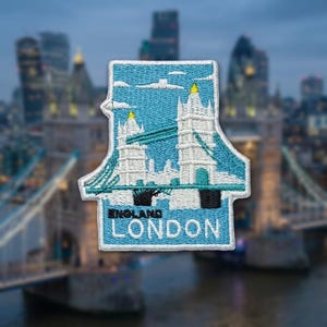 May include: A rectangular embroidered patch featuring a light blue sky with white clouds above a white and teal depiction of Tower Bridge. The text ENGLAND and LONDON is stitched in black and white lettering at the bottom of the patch.