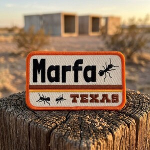 May include: A rectangular embroidered patch with an orange border. The white background features the black text Marfa, a black ant illustration, horizontal brown and yellow stripes, and the brown text TEXAS with two small black ants.
