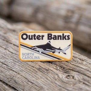 May include: A rectangular embroidered patch with a cream background and a yellow border. It features the black text Outer Banks and light blue text North Carolina. A black and white shark graphic is positioned over diagonal yellow and grey lines.