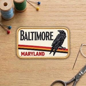 May include: Embroidered patch featuring a black raven perched on a branch, with the word "BALTIMORE" above and "MARYLAND" below. The patch has a cream background with red, orange, and black stripes. Sewing supplies are in the background.