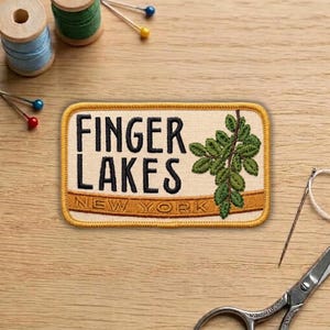 May include: Embroidered patch with the words "FINGER LAKES NEW YORK" in black lettering. A green leaf design is on the right side. The patch has a tan background with a gold border. Sewing supplies are in the background.