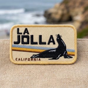 May include: A rectangular embroidered patch with a beige background and a yellow border. It features a black sea lion, the text LA JOLLA in black, and the word CALIFORNIA in smaller black letters, with horizontal blue and gold stripes.